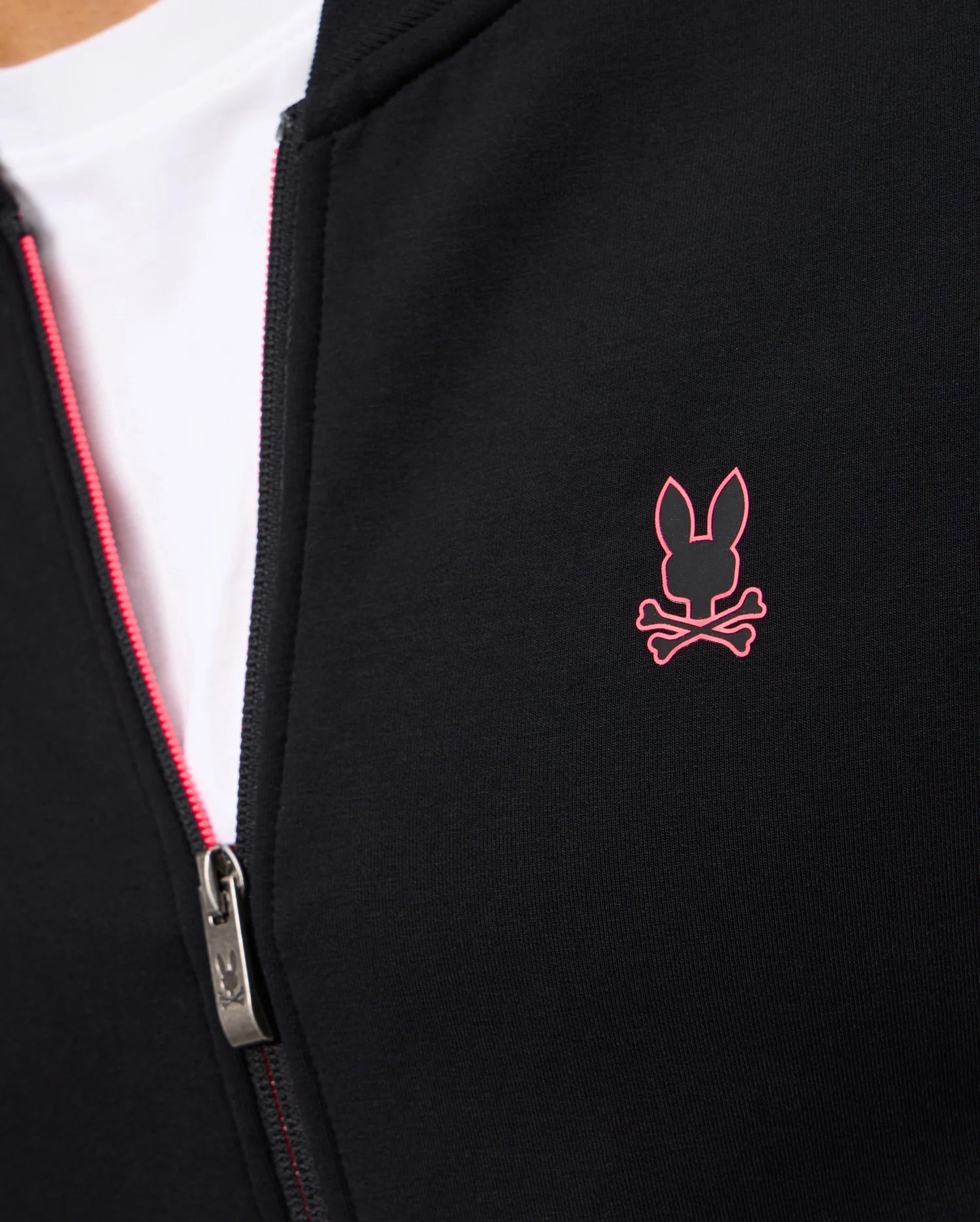 Close-up of a person wearing a black Psycho Bunny MENS TRAVIS TRACK JACKET (B6J142D200) with a white shirt underneath. The jacket features a red zipper and a small red bunny logo with crossed bones on the chest, along with invisible zip pockets.
