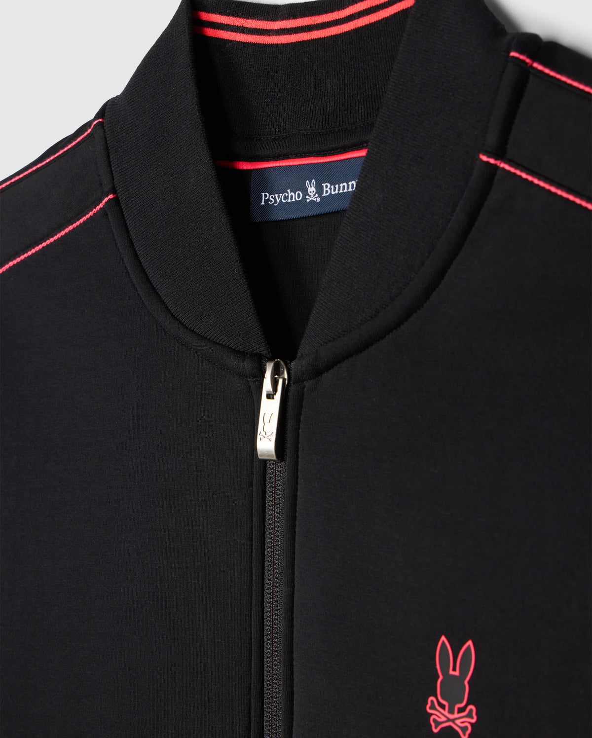 Close-up of the Psycho Bunny MENS TRAVIS TRACK JACKET - B6J142D200 in black with red detailing. The collar and shoulders feature thin red stripes, and a small embroidered bunny logo with crossbones appears on the left side of the chest. Invisible zip pockets add functionality to this stylish piece.
