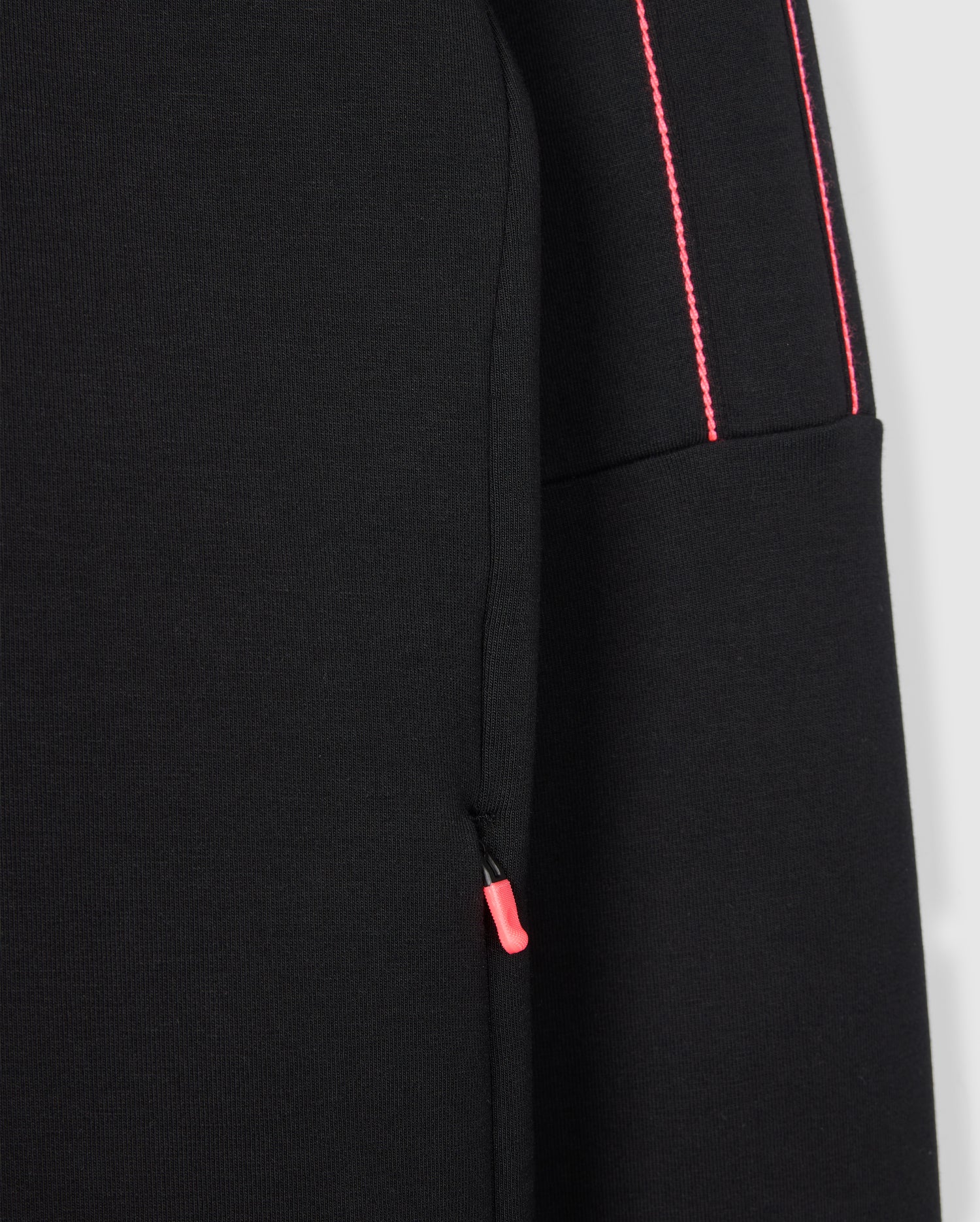 Close-up of a Psycho Bunny MENS TRAVIS TRACK JACKET - B6J142D200 in black. It features a pocket with a red zipper pull tab and two parallel red stitches running vertically along the seam. The fabric appears to be smooth and structured, making it perfect for transitioning from training to recovery.