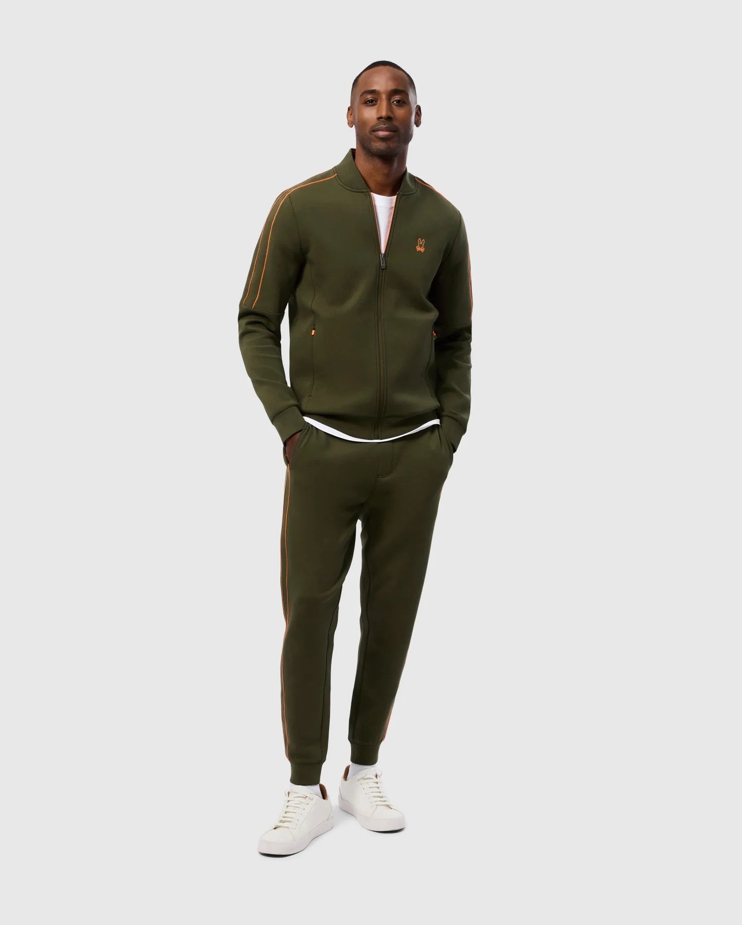 A man stands confidently in his Psycho Bunny Mens Travis Track Jacket - B6J142D200 and matching set, crafted from high-quality cotton-blend fabric. The olive-green ensemble features zipper and stripe details along the arms and legs. He pairs it with a white t-shirt underneath and white sneakers against a plain light gray background.