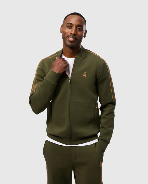 A person is smiling at the camera while wearing a high-quality cotton-blend olive green Psycho Bunny MENS TRAVIS TRACK JACKET (B6J142D200) with orange piping, part of a matching set. The tracksuit top is zipped up halfway, revealing a white shirt underneath. They have one hand in their pocket and are posing against a plain, light grey background.
