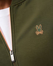 Close-up of a person wearing a green MENS TRAVIS TRACK JACKET - B6J142D200 from Psycho Bunny over a white shirt. The high-quality cotton-blend jacket features a small orange bunny head with crossbones logo on the chest and an X and O design on the zipper pull, perfect for pairing with the matching set.