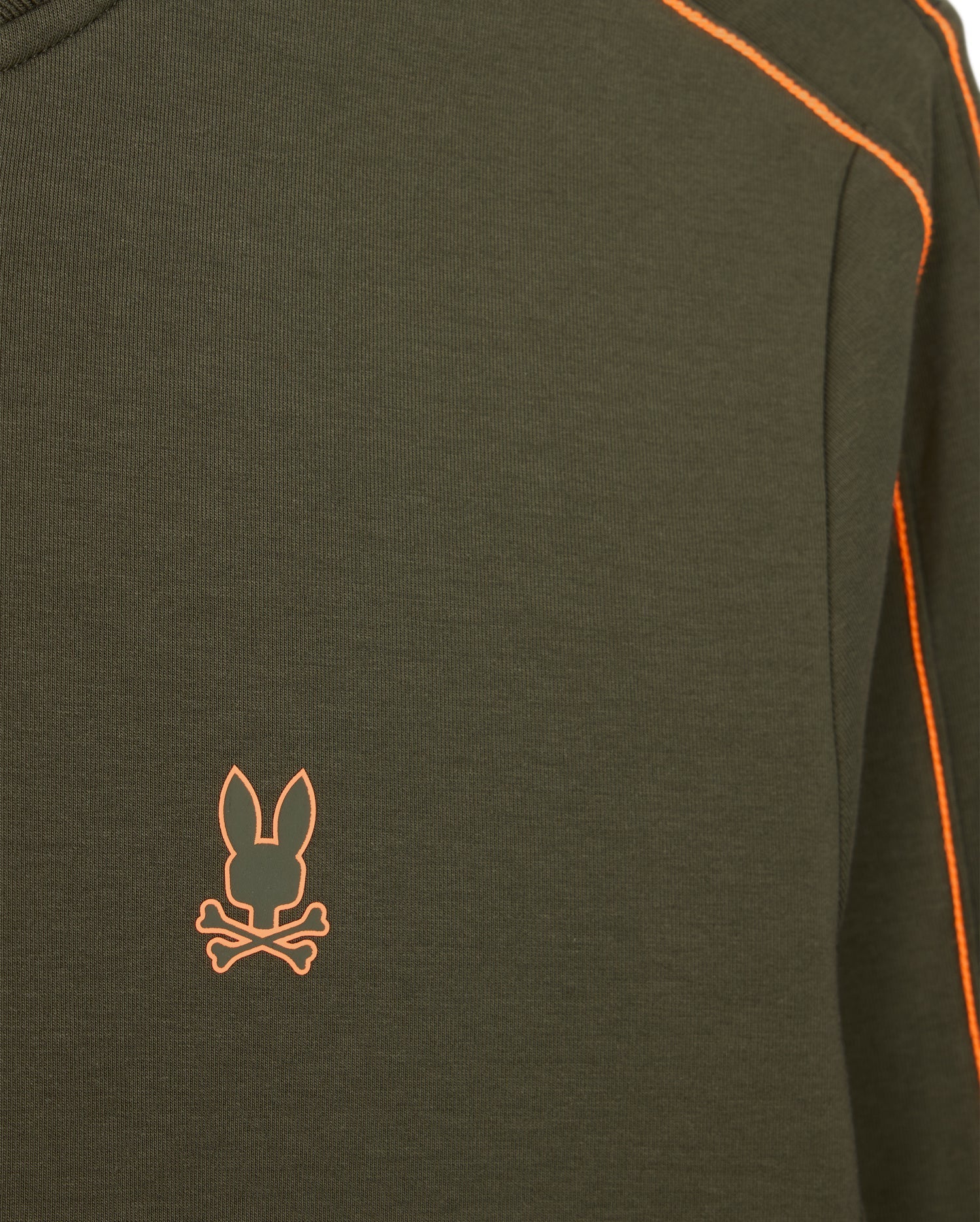 Close-up of the Psycho Bunny MENS TRAVIS TRACK JACKET - B6J142D200, a dark green high-quality cotton-blend garment with orange stitching along the seams. This jacket, part of a matching set, features an orange emblem on the left chest area depicting a rabbit head above crossed bones.