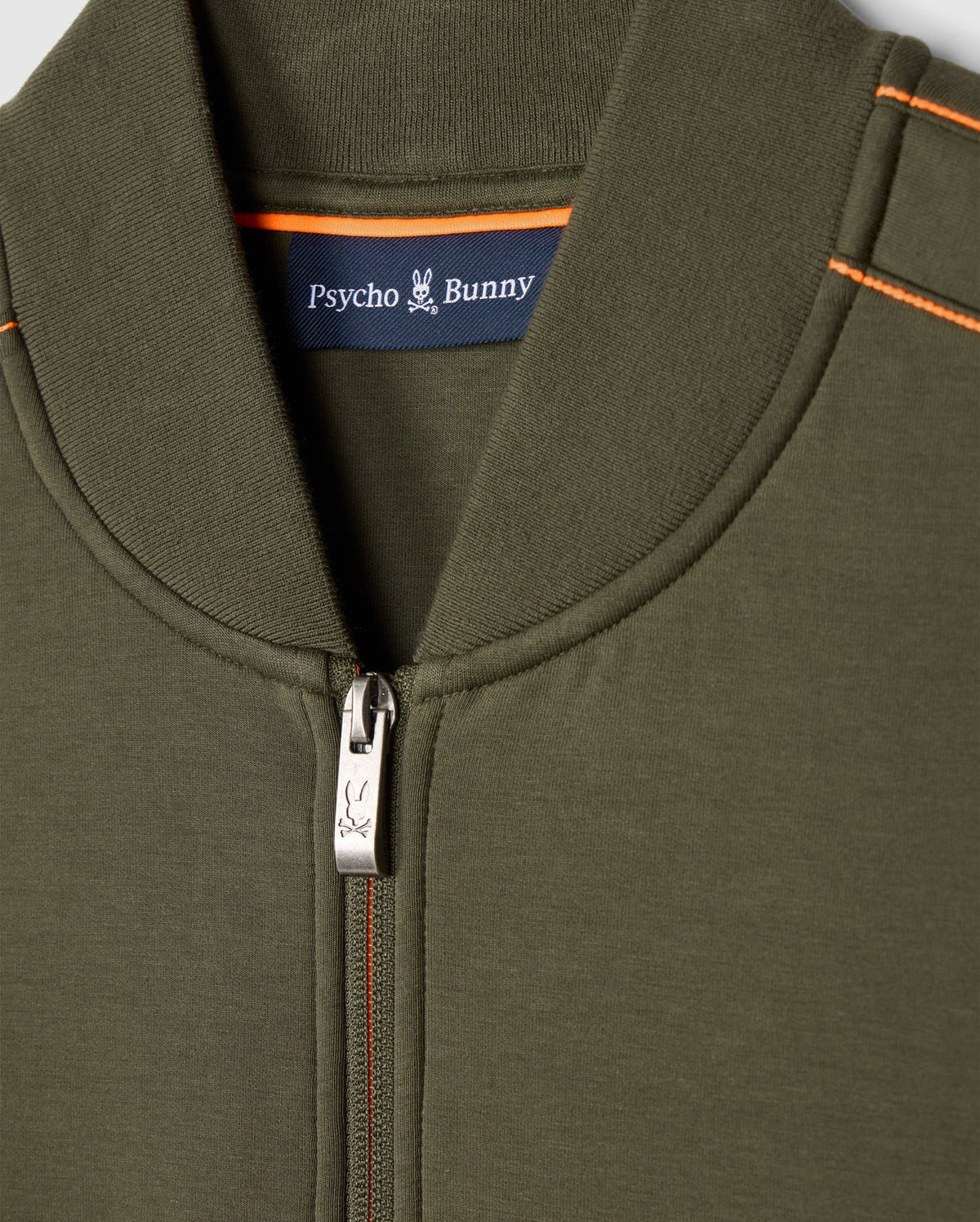 Close-up of the Psycho Bunny men's Travis track jacket in olive green (B6J142D200), featuring a zipper with a bunny logo on the pull. The inner collar displays a label with "Psycho Bunny" and a small bunny logo. Made from high-quality cotton-blend, this jacket includes orange stitching on the shoulders.