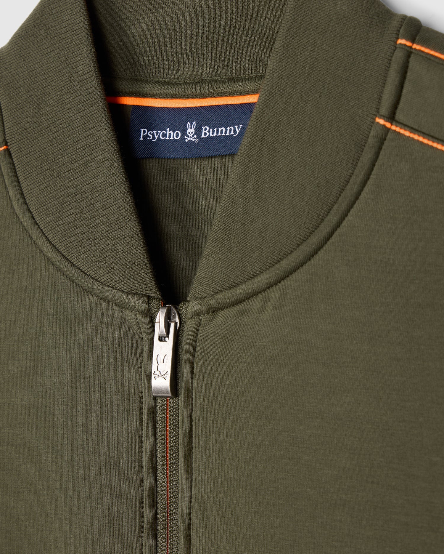 Close-up of the Psycho Bunny men's Travis track jacket in olive green (B6J142D200), featuring a zipper with a bunny logo on the pull. The inner collar displays a label with "Psycho Bunny" and a small bunny logo. Made from high-quality cotton-blend, this jacket includes orange stitching on the shoulders.