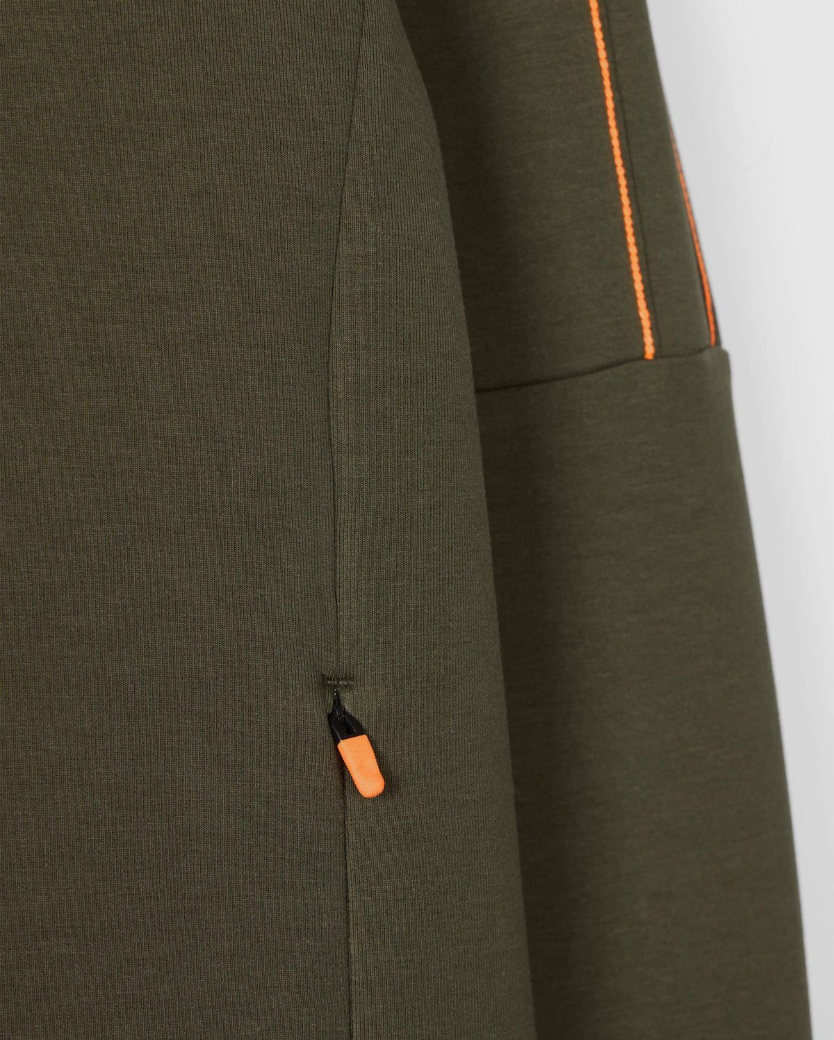 Close-up of the olive green Psycho Bunny Men's Travis Track Jacket (B6J142D200) featuring an orange pull tab on a zippered pocket. Orange stitching detail is visible along a separate seam, contrasting beautifully with the high-quality cotton-blend fabric's texture.