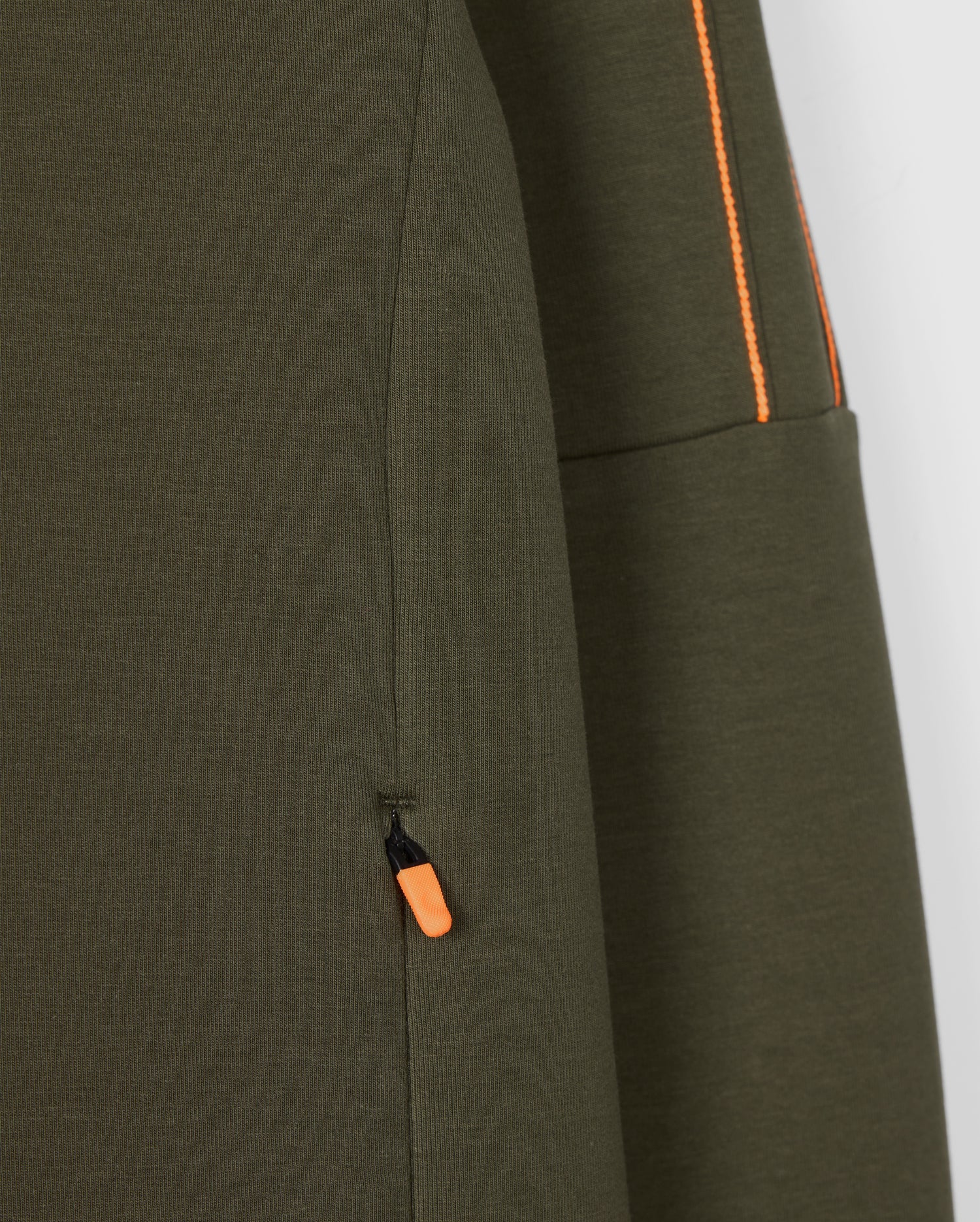 Close-up of the olive green Psycho Bunny Men's Travis Track Jacket (B6J142D200) featuring an orange pull tab on a zippered pocket. Orange stitching detail is visible along a separate seam, contrasting beautifully with the high-quality cotton-blend fabric's texture.
