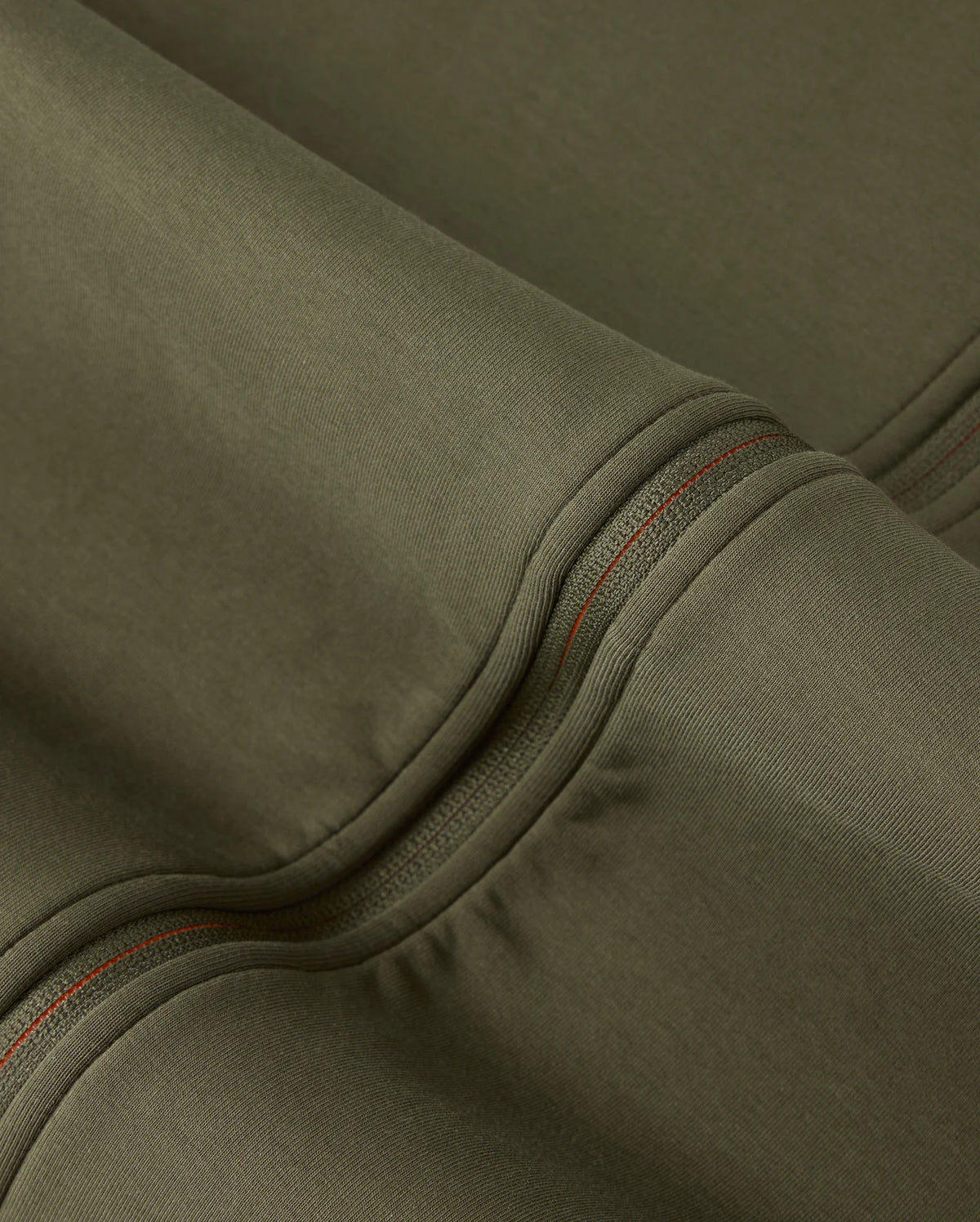Close-up of a piece of olive green, high-quality cotton-blend fabric showcasing detailed stitching with red thread. The material appears smooth and sturdy, with prominent seams and folds creating subtle shadows and highlights, reminiscent of the craftsmanship found in the Mens Travis Track Jacket (B6J142D200) by Psycho Bunny.