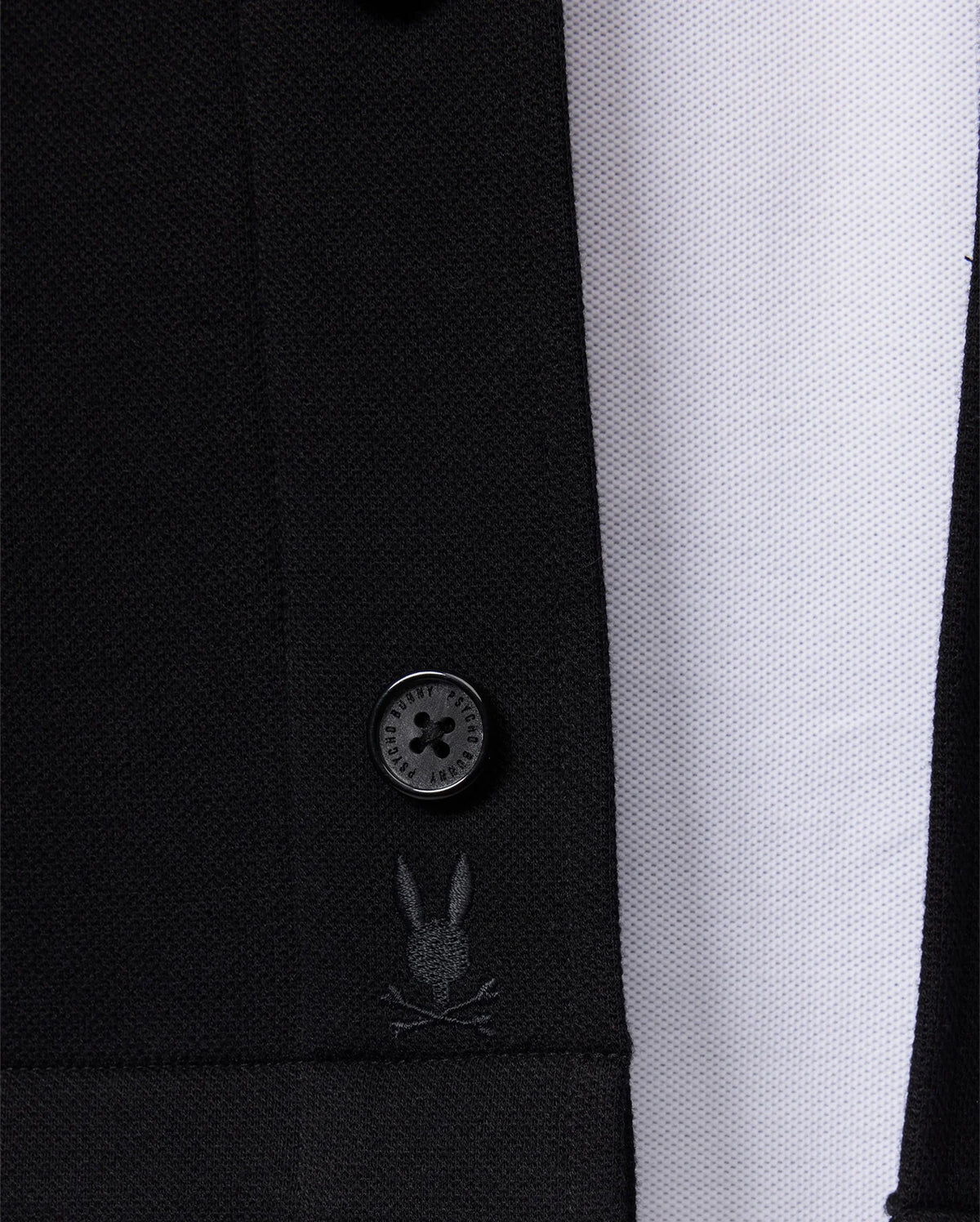 Close-up of the Psycho Bunny Men's Bedford Shirt Jacket (B6J290B200) showcasing a prominently visible button and an embroidered design featuring a small rabbit face with crossbones. The work-jacket style's texture stands out beautifully against a white, textured background.
