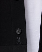 Close-up of the Psycho Bunny Men's Bedford Shirt Jacket (B6J290B200) showcasing a prominently visible button and an embroidered design featuring a small rabbit face with crossbones. The work-jacket style's texture stands out beautifully against a white, textured background.