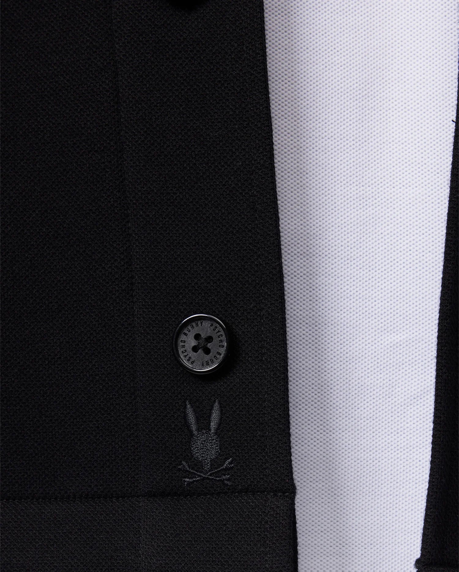 Close-up of the Psycho Bunny Men's Bedford Shirt Jacket (B6J290B200) showcasing a prominently visible button and an embroidered design featuring a small rabbit face with crossbones. The work-jacket style's texture stands out beautifully against a white, textured background.