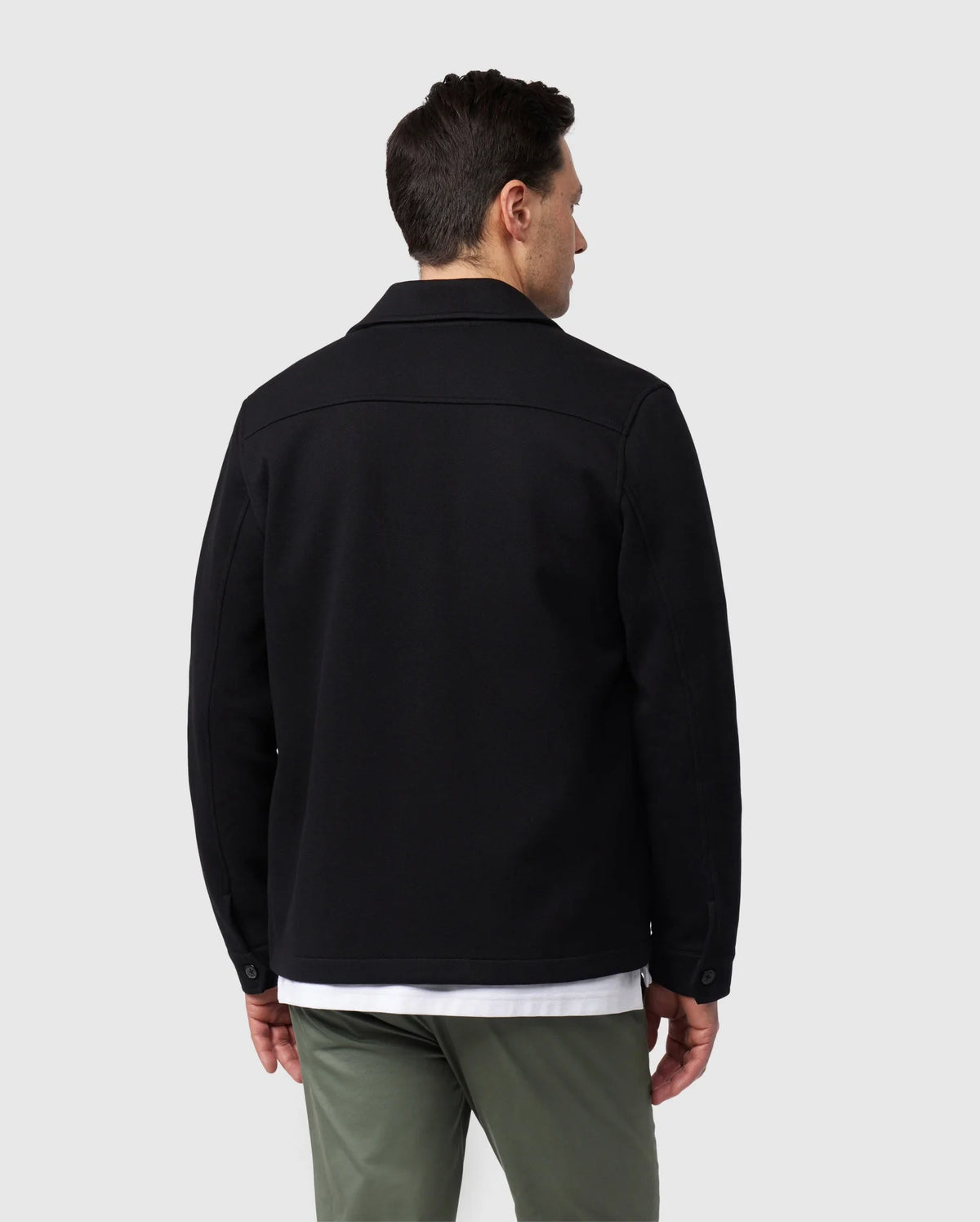 A man stands with his back to the camera, wearing a fashionable jacket ensemble from Psycho Bunny. He sports the MENS BEDFORD SHIRT JACKET (B6J290B200) in black, paired with a clean white shirt and green pants. The understated gray background highlights the timeless work-jacket style of his outfit.