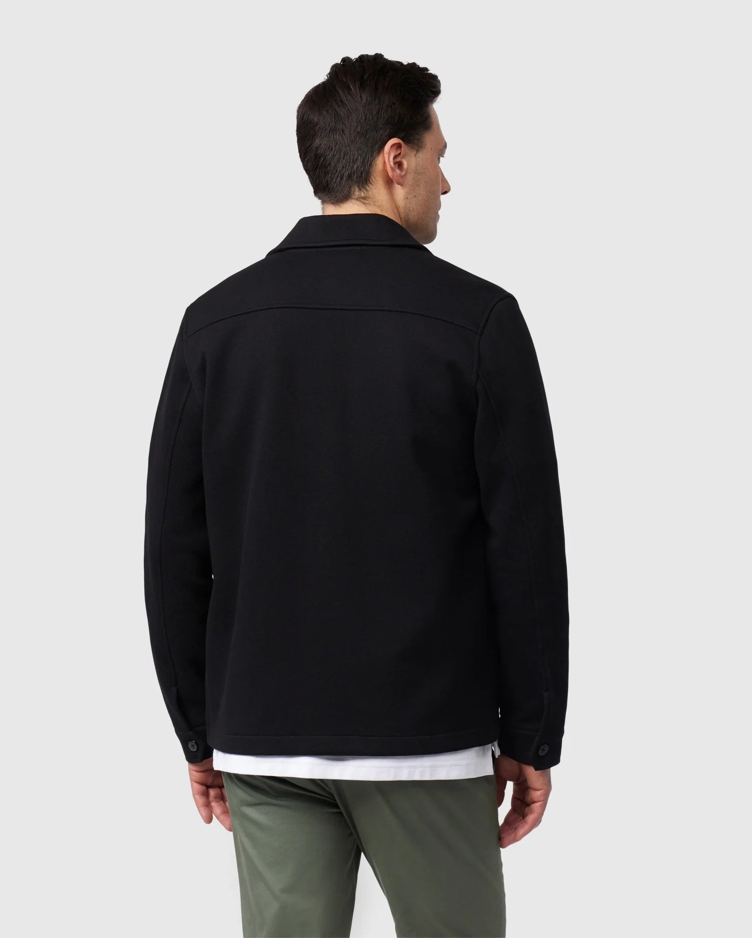 A man stands with his back to the camera, wearing a fashionable jacket ensemble from Psycho Bunny. He sports the MENS BEDFORD SHIRT JACKET (B6J290B200) in black, paired with a clean white shirt and green pants. The understated gray background highlights the timeless work-jacket style of his outfit.