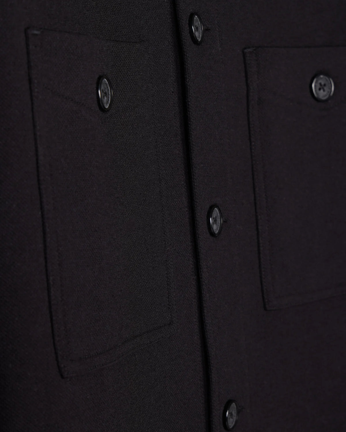 Close-up of the Psycho Bunny Men's Bedford Shirt Jacket - B6J290B200 in black fabric, showcasing two pockets and a matching row of buttons. The textured material evokes a work-jacket style, with pockets featuring button closures.