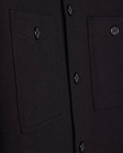 Close-up of the Psycho Bunny Men's Bedford Shirt Jacket - B6J290B200 in black fabric, showcasing two pockets and a matching row of buttons. The textured material evokes a work-jacket style, with pockets featuring button closures.