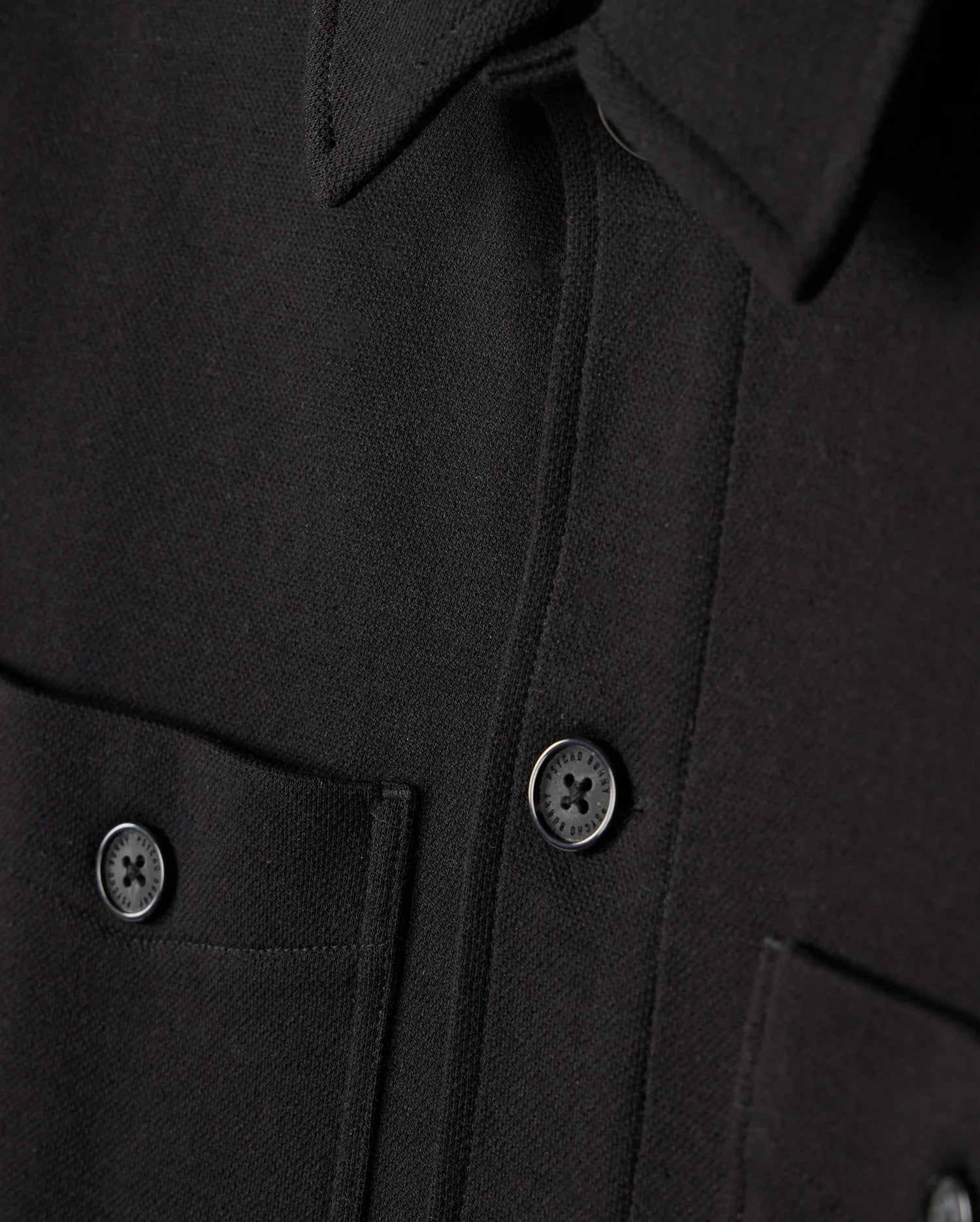 Close-up of the Psycho Bunny MENS BEDFORD SHIRT JACKET - B6J290B200 in black, showcasing two dark buttons and dual chest pockets. The fabric exhibits a textured appearance analogous to work-jacket style, with the men's jacket neatly buttoned.
