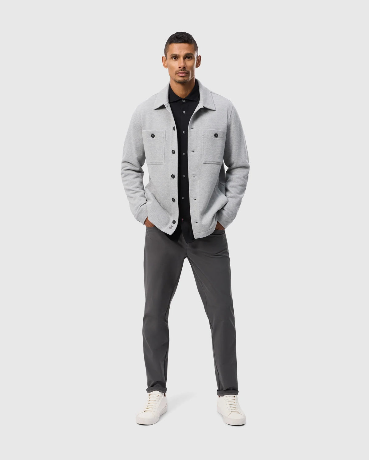 A man confidently poses against a plain background, wearing the Psycho Bunny BEDFORD SHIRT JACKET - B6J290B200 in light gray over a black shirt, paired with dark gray pants and white sneakers. His hands are tucked into his jacket pockets as he stands facing forward.