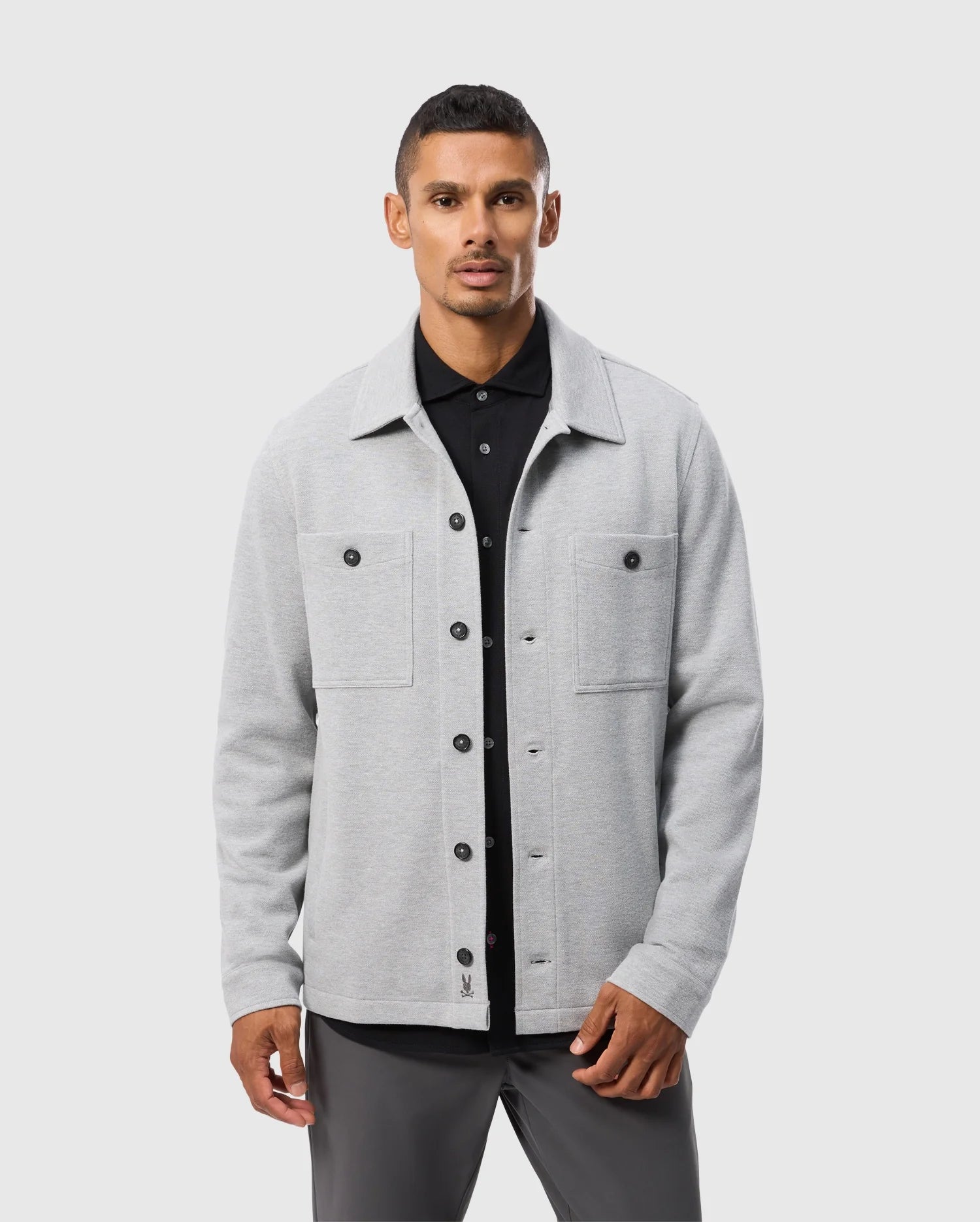 A man stands against a plain background wearing the Psycho Bunny BEDFORD SHIRT JACKET (B6J290B200), a light grey button-up with two chest pockets, over a black shirt. He looks forward with a neutral expression.