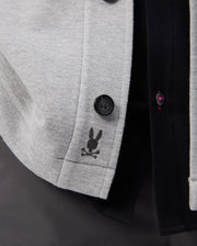 A close-up of the Psycho Bunny BEDFORD SHIRT JACKET - B6J290B200 in gray fabric highlights a rabbit head and crossbones logo, with black and dark red buttons against a dark background.