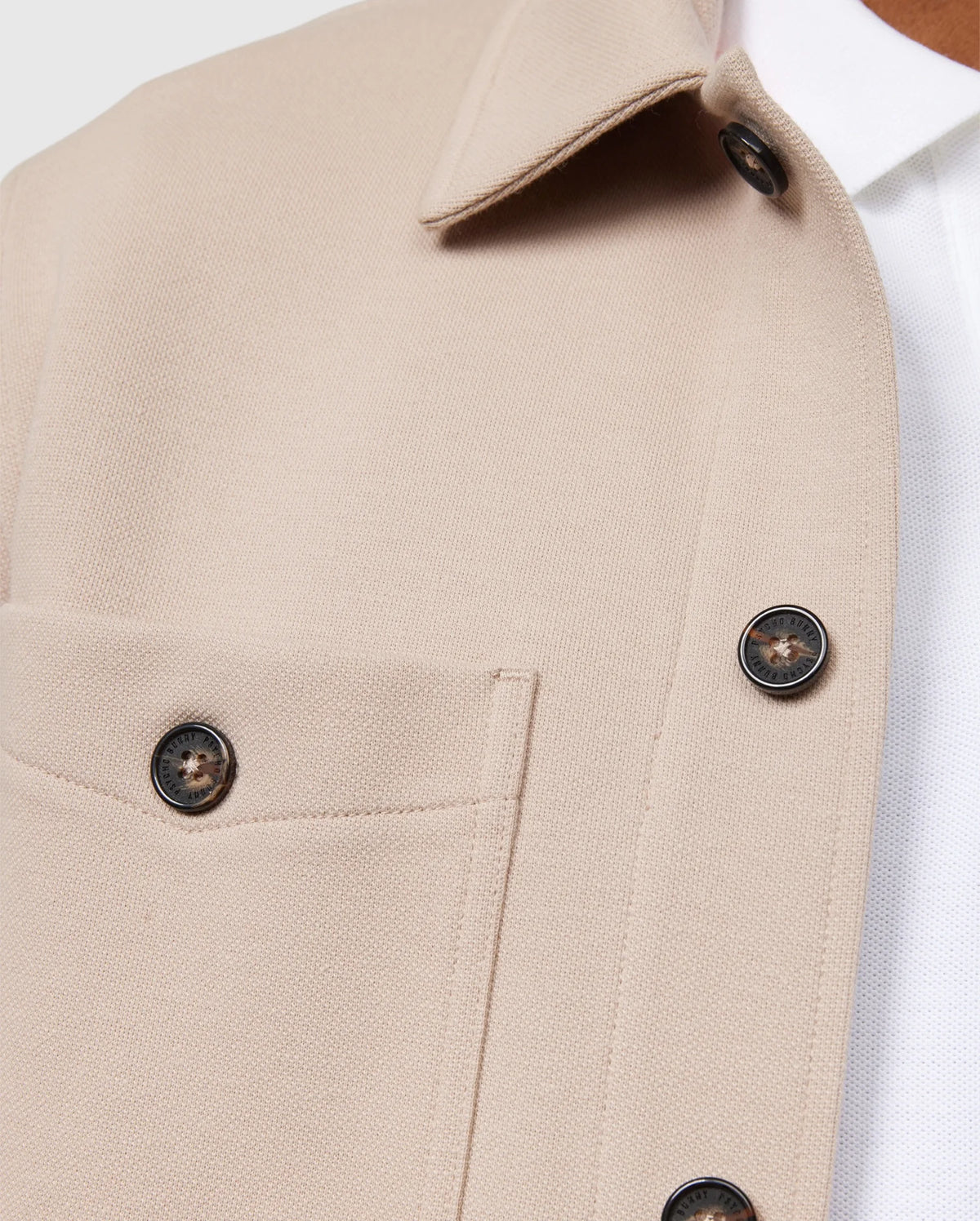 Close-up of the Psycho Bunny MENS BEDFORD SHIRT JACKET - B6J290B200 in beige, showcasing logo-engraved dark buttons and work-jacket style pockets, layered over a white shirt. The textured fabric and focus on the upper part highlight its structured design akin to a classic Bedford shirt jacket.