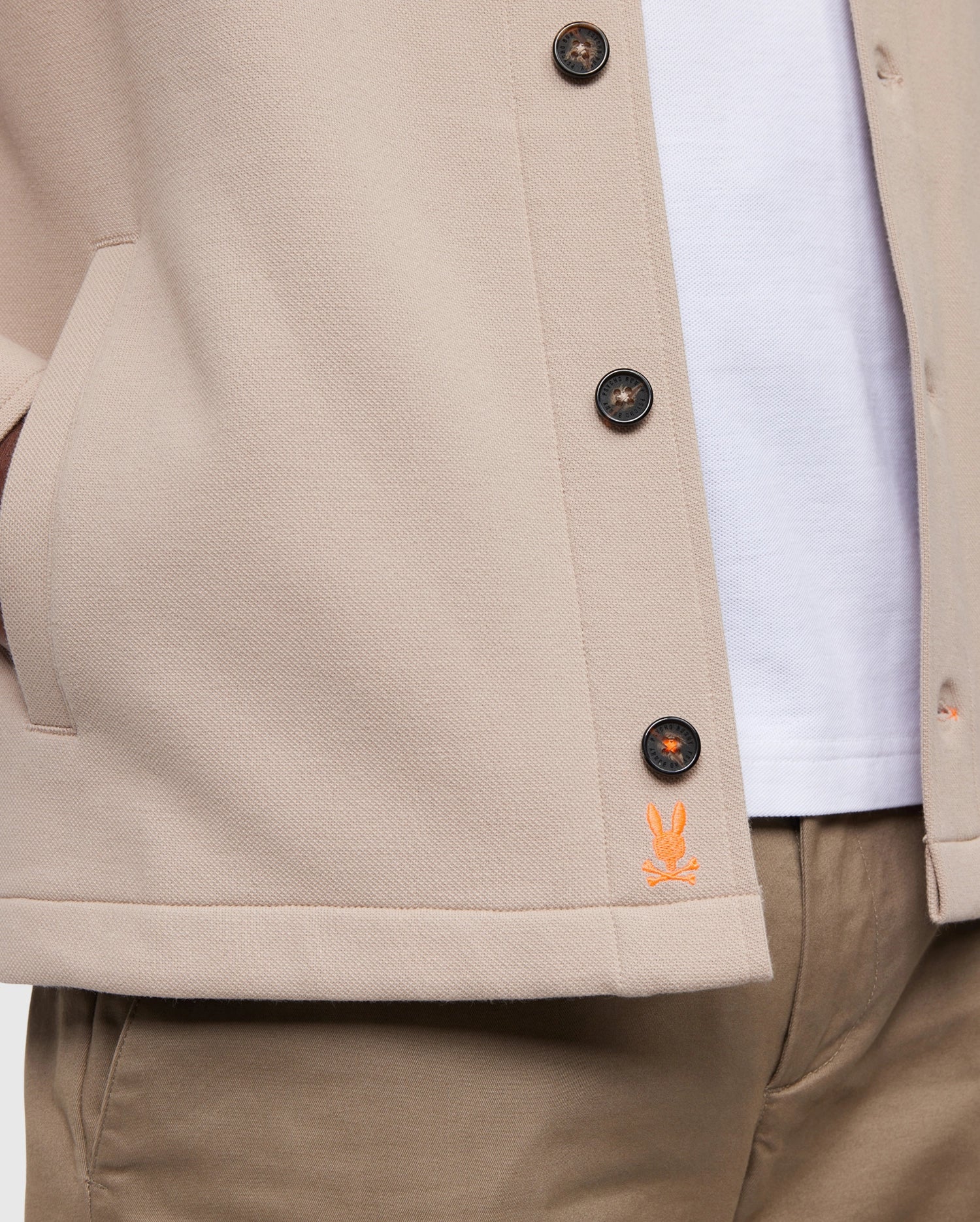 Close-up of a person wearing the light beige Psycho Bunny MENS BEDFORD SHIRT JACKET - B6J290B200, showcasing logo-engraved black buttons and an orange embroidered logo near the hem. The work-jacket style pockets hold their hands, paired elegantly with a white shirt and khaki pants.