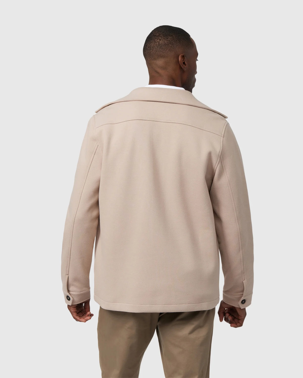 A man stands with his back to the camera, dressed in a beige MENS BEDFORD SHIRT JACKET - B6J290B200 from Psycho Bunny, featuring work-jacket style pockets, paired with khaki pants against a plain gray background.