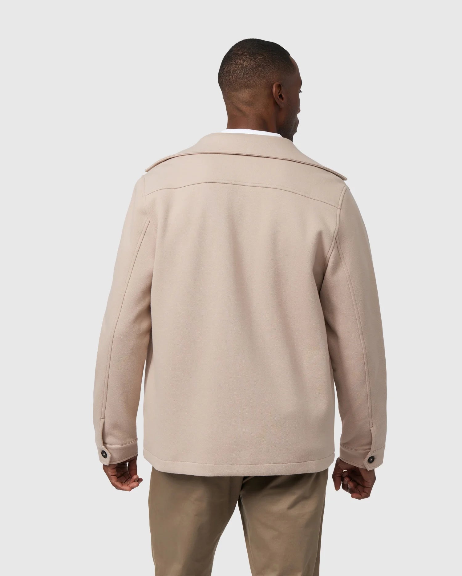 A man stands with his back to the camera, dressed in a beige MENS BEDFORD SHIRT JACKET - B6J290B200 from Psycho Bunny, featuring work-jacket style pockets, paired with khaki pants against a plain gray background.