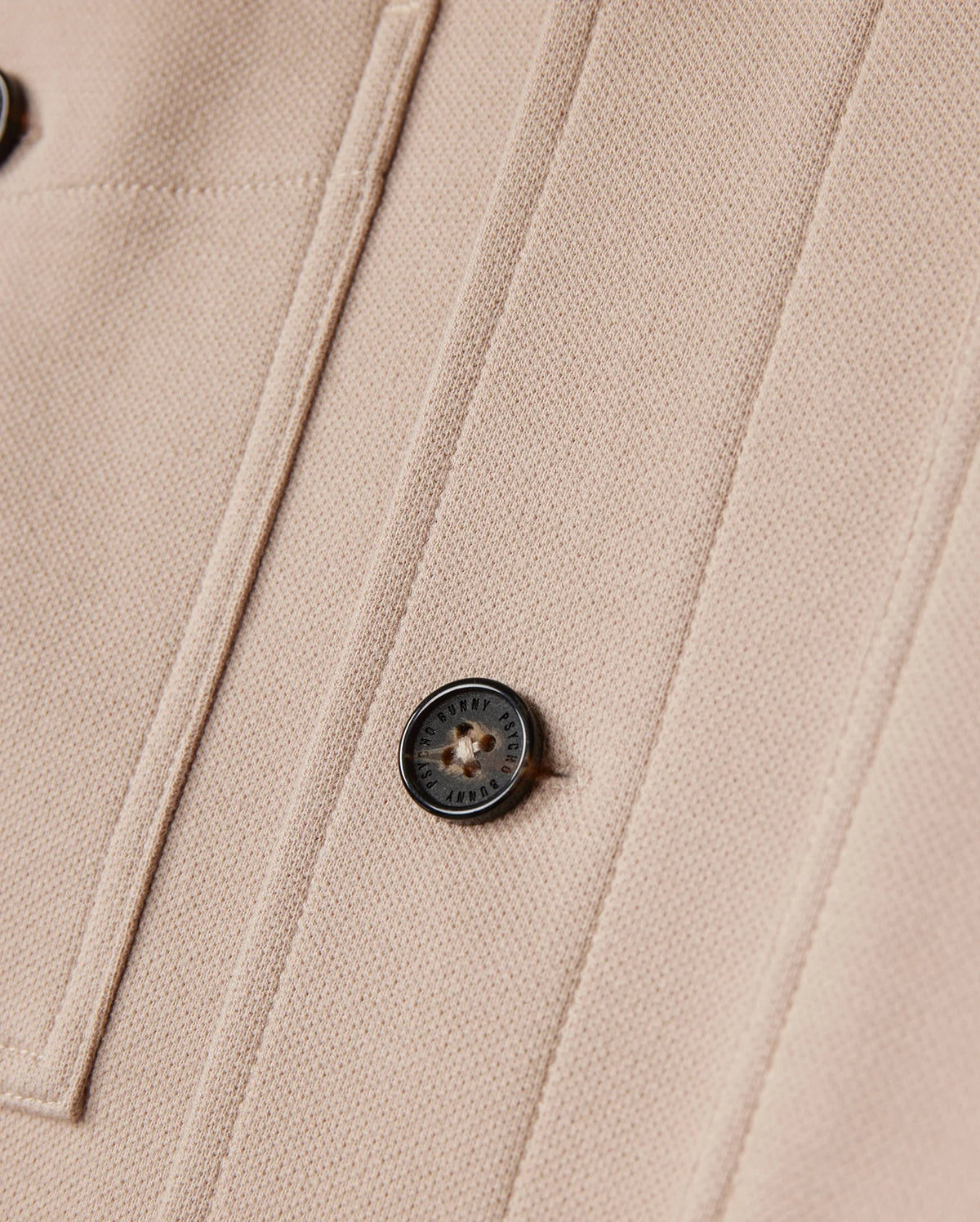 Close-up of a beige fabric from the Psycho Bunny MENS BEDFORD SHIRT JACKET - B6J290B200, showcasing a black button engraved with the logo and text, alongside parallel seams forming a rectangular pattern. The textured fabric suggests it's part of an item such as this shirt jacket.