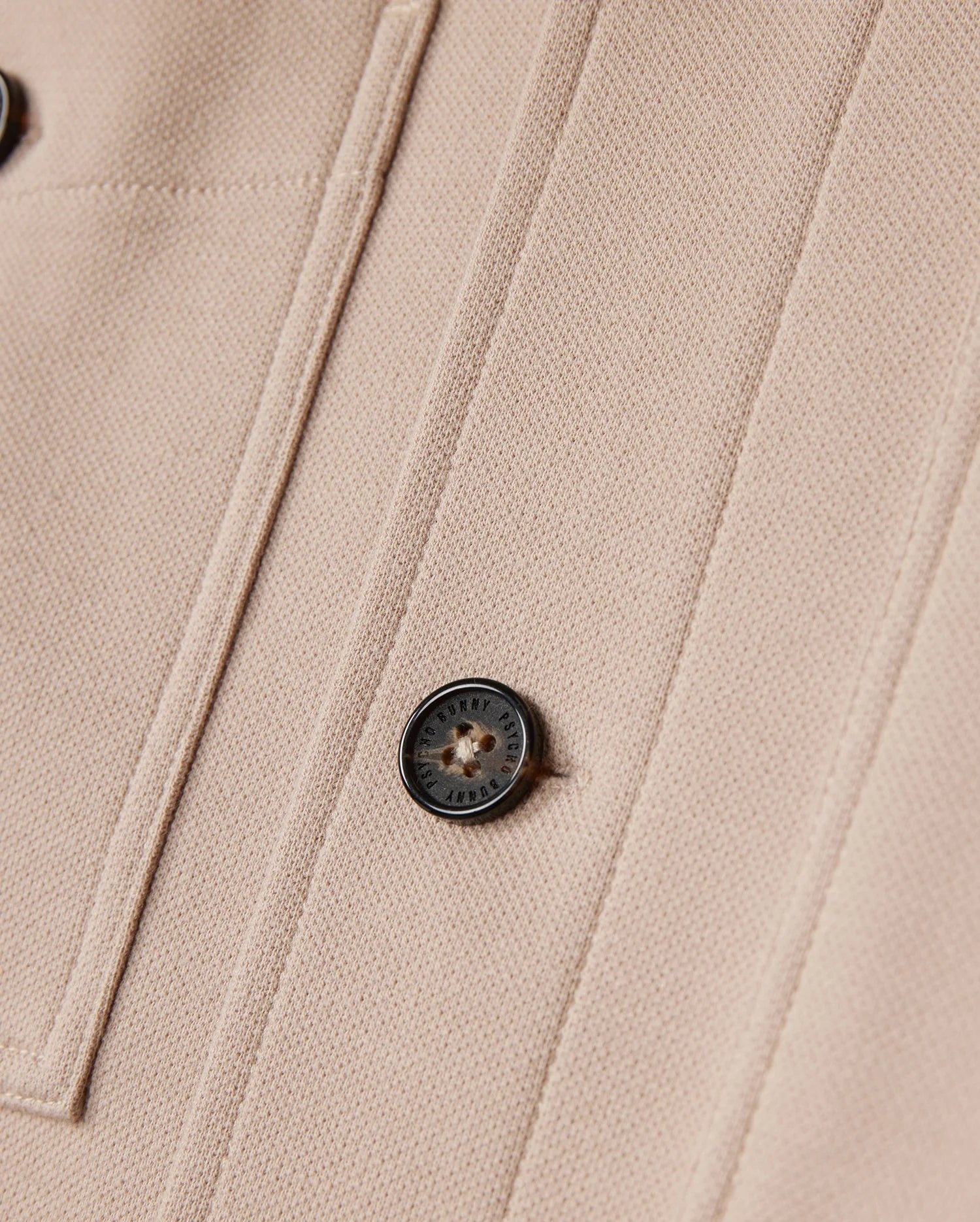Close-up of a beige fabric from the Psycho Bunny MENS BEDFORD SHIRT JACKET - B6J290B200, showcasing a black button engraved with the logo and text, alongside parallel seams forming a rectangular pattern. The textured fabric suggests it's part of an item such as this shirt jacket.