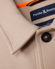 Close-up of the Psycho Bunny beige collared MENS BEDFORD SHIRT JACKET - B6J290B200, featuring a black button engraved with the logo. The neck is adorned with a "Psycho Bunny" label and a small bunny logo. The textured material resembles that of a Bedford shirt jacket, exuding subtle sophistication.