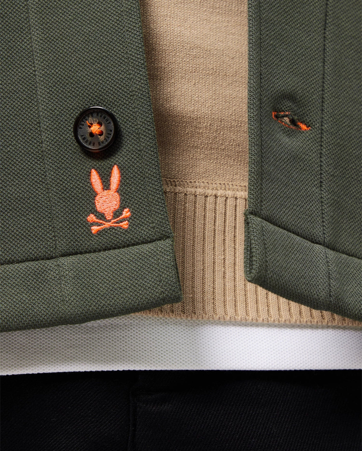 Close-up of the MENS BEDFORD SHIRT JACKET - B6J290B200 by Psycho Bunny, featuring a green cotton-blend fabric and an orange embroidered logo depicting a rabbit skull and crossbones. The urban vibe is accentuated as the jacket is styled open, showcasing a beige sweater underneath.