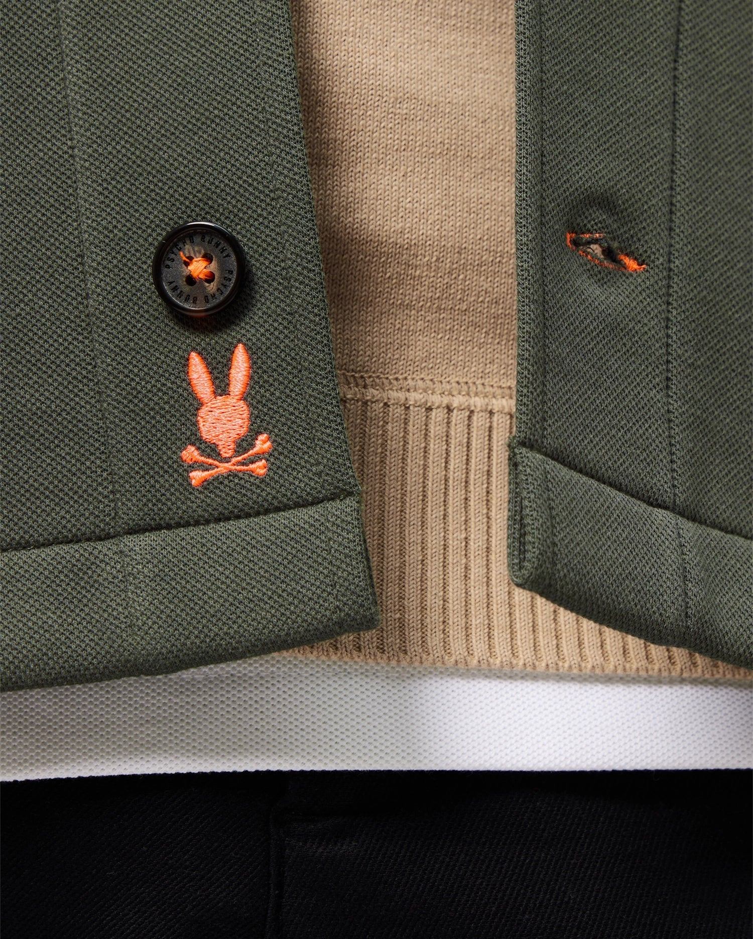 Close-up of the MENS BEDFORD SHIRT JACKET - B6J290B200 by Psycho Bunny, featuring a green cotton-blend fabric and an orange embroidered logo depicting a rabbit skull and crossbones. The urban vibe is accentuated as the jacket is styled open, showcasing a beige sweater underneath.