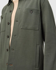 Close-up of a person wearing the Psycho Bunny MENS BEDFORD SHIRT JACKET - B6J290B200 in dark green over a beige shirt. This cotton-blend overshirt, with its urban vibe, showcases two chest pockets and black buttons.