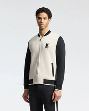 A man models the Psycho Bunny MENS CRETE COLOR BLOCK BOMBER (B6J421F200) with a bunny logo, featuring ribbed cuffs and a zip closure. Paired with black pants against a light gray background, this classic cut jacket is an ideal transitional wardrobe choice.