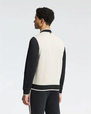 A person dons the Psycho Bunny MENS CRETE COLOR BLOCK BOMBER - B6J421F200 with black sleeves and a white back, paired with matching pants featuring a white stripe. The sleek silhouette stands against a plain background, facing away from the camera.