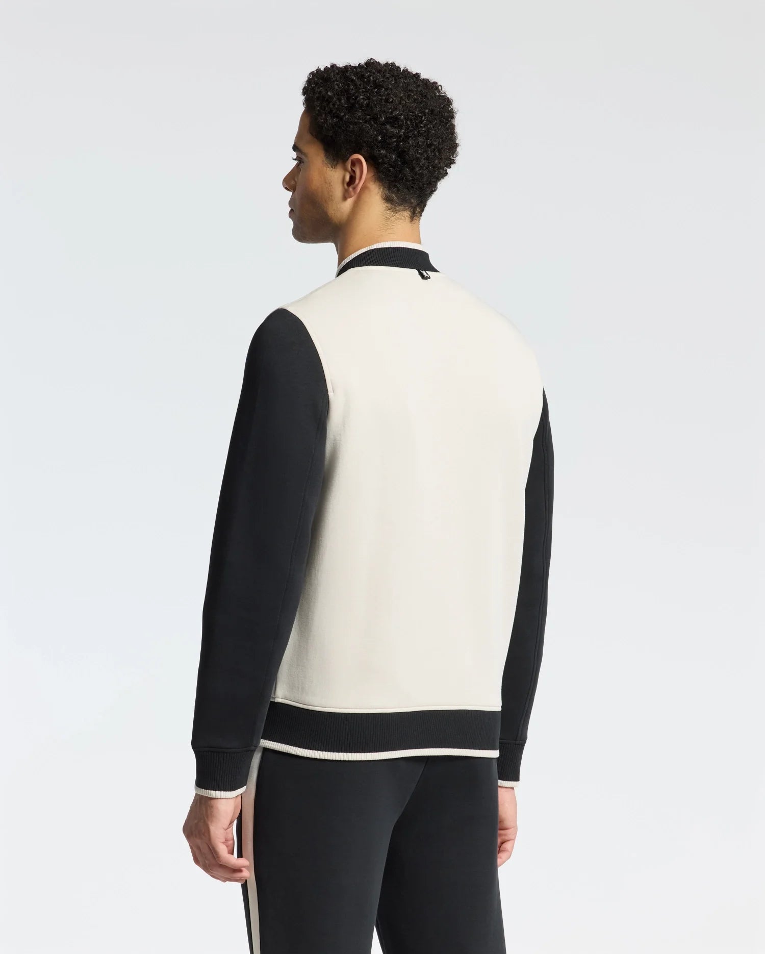 A person dons the Psycho Bunny MENS CRETE COLOR BLOCK BOMBER - B6J421F200 with black sleeves and a white back, paired with matching pants featuring a white stripe. The sleek silhouette stands against a plain background, facing away from the camera.