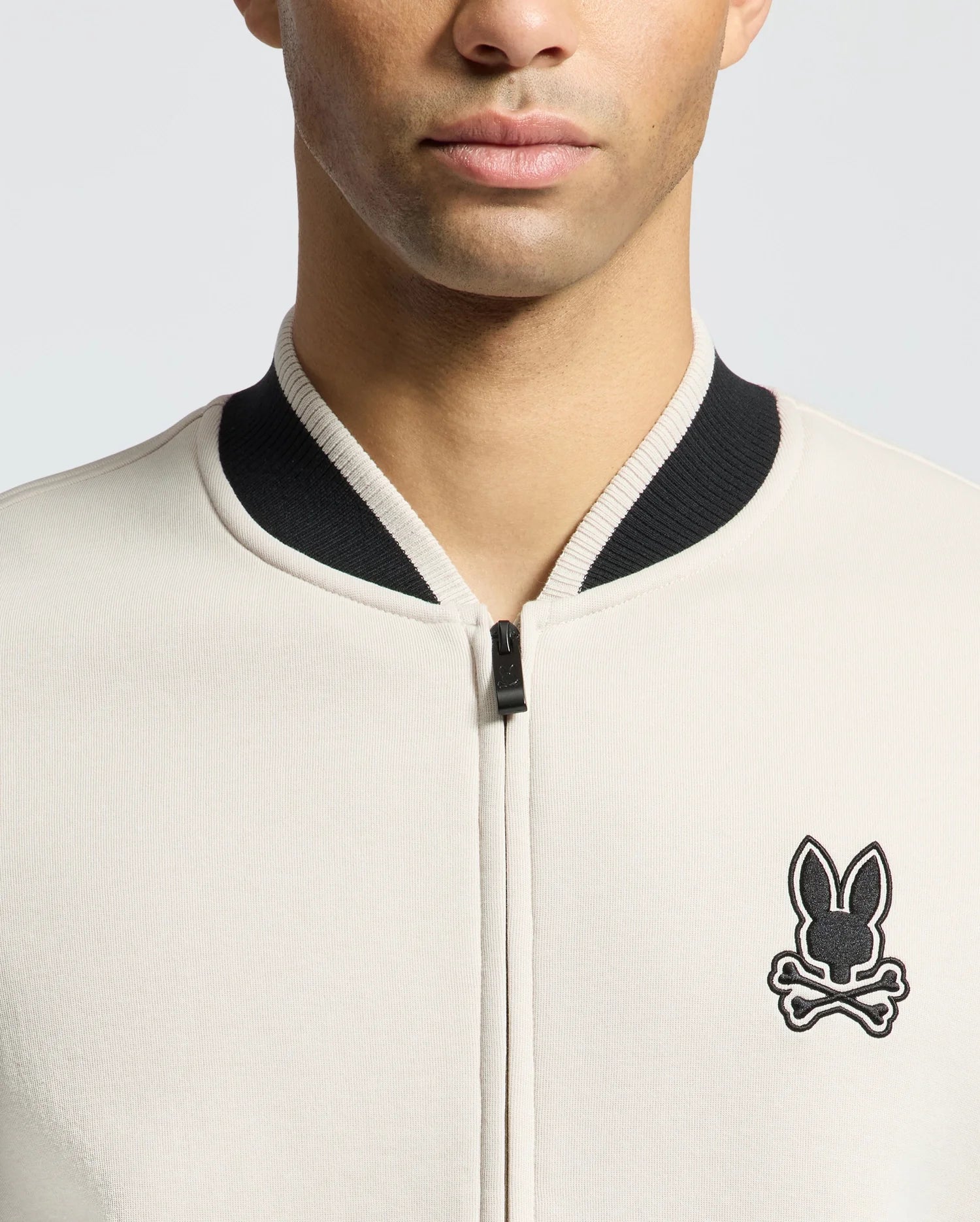 Wearing the Psycho Bunny MENS CRETE COLOR BLOCK BOMBER - B6J421F200, a person sports a classic light beige zip-up jacket with a black collar and stylized bunny emblem. The image captures them against a gray backdrop, face partially visible and cropped at the eyes.
