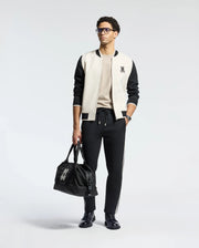 A person stylishly pairs the Psycho Bunny MENS CRETE COLOR BLOCK BOMBER - B6J421F200 featuring a rabbit logo with a beige shirt and black joggers with white stripes as a perfect transitional outfit. Black shoes and a matching logo bag complete the look against a plain background.