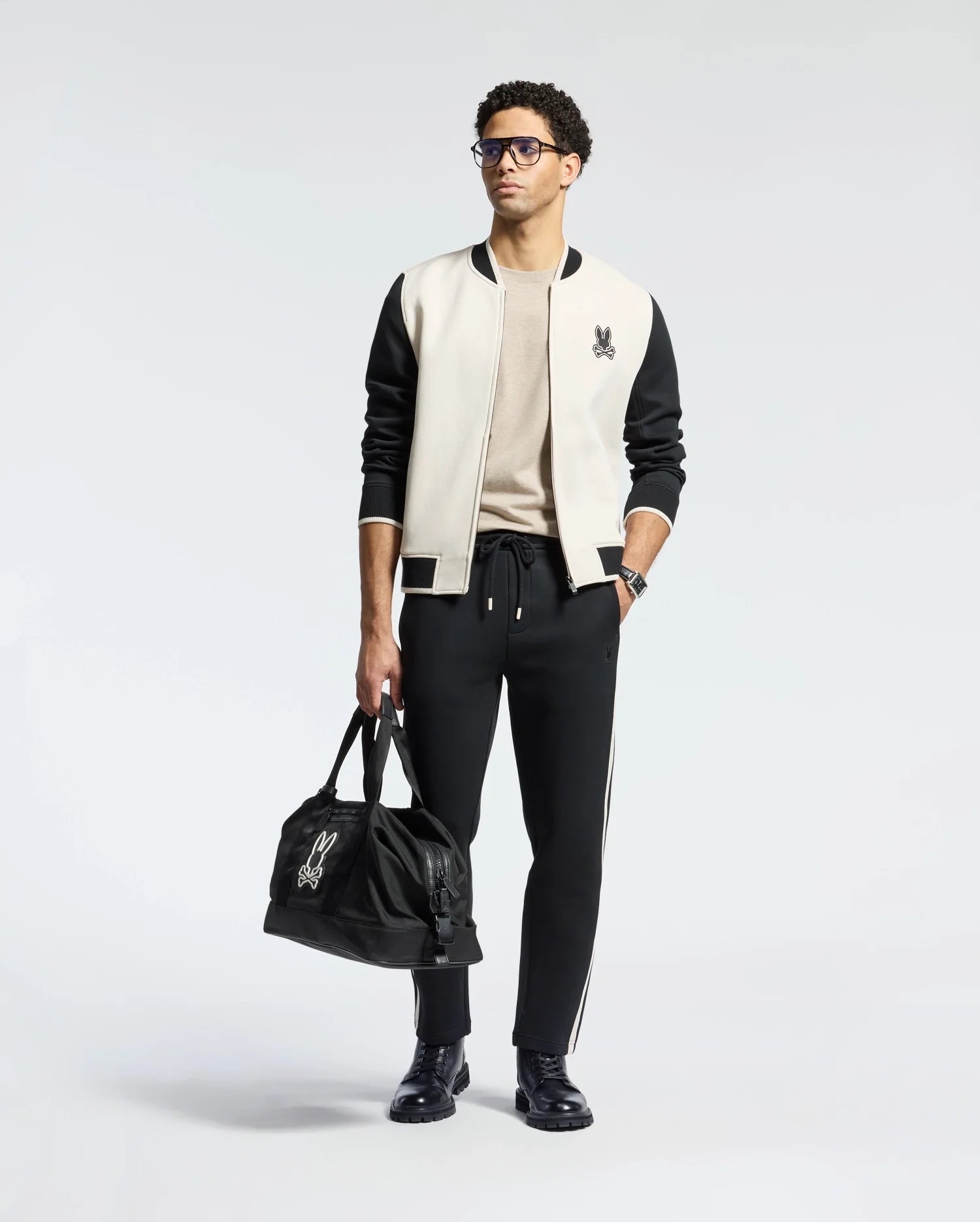 A person stylishly pairs the Psycho Bunny MENS CRETE COLOR BLOCK BOMBER - B6J421F200 featuring a rabbit logo with a beige shirt and black joggers with white stripes as a perfect transitional outfit. Black shoes and a matching logo bag complete the look against a plain background.