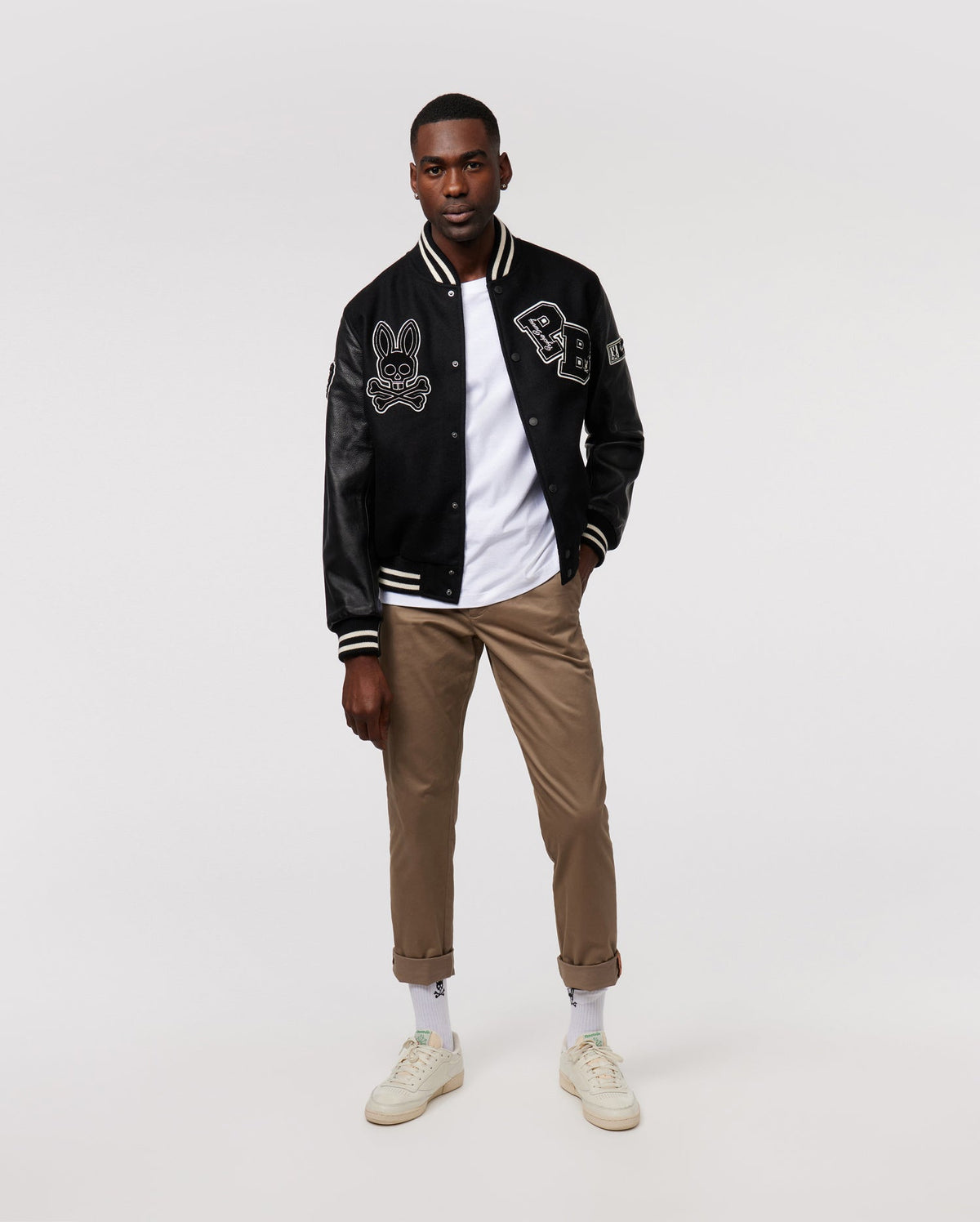 A man stands confidently wearing the Psycho Bunny MENS ROBBINS VARSITY BOMBER JACKET - B6J524Z1OW, along with a white t-shirt, khaki pants, and white sneakers. His hands rest in his pockets as he faces forward against a plain, light background.