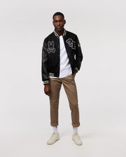 A man stands confidently wearing the Psycho Bunny MENS ROBBINS VARSITY BOMBER JACKET - B6J524Z1OW, along with a white t-shirt, khaki pants, and white sneakers. His hands rest in his pockets as he faces forward against a plain, light background.