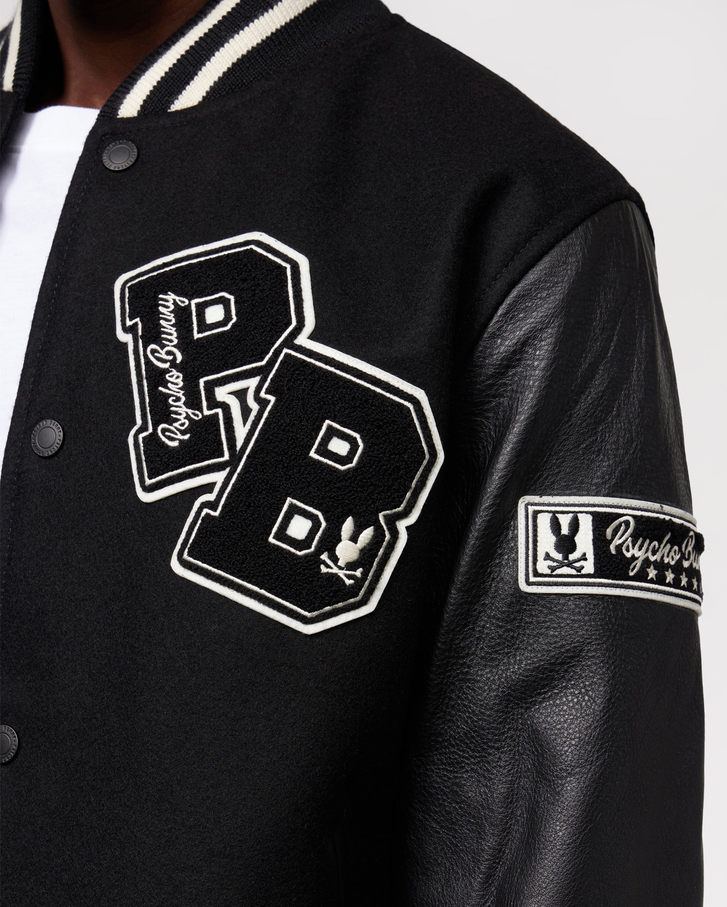 A close-up captures someone in the Men's Robbins Varsity Bomber Jacket - B6J524Z1OW by Psycho Bunny, showcasing its black wool-blend body and smooth black leather sleeves. White "PB" chenille patches and "Psycho Bunny" text stand out on the fabric, while a distinctive patch with a bunny and stars adorns the arm.