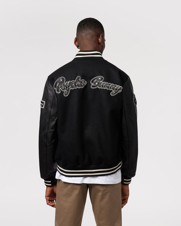 A person stands with their back to the camera, wearing the MENS ROBBINS VARSITY BOMBER JACKET - B6J524Z1OW by Psycho Bunny. The black jacket is adorned with chenille patches and features "Psycho Bunny" written across the back in white script. They also wear khaki pants and a white shirt beneath this stylish jacket.