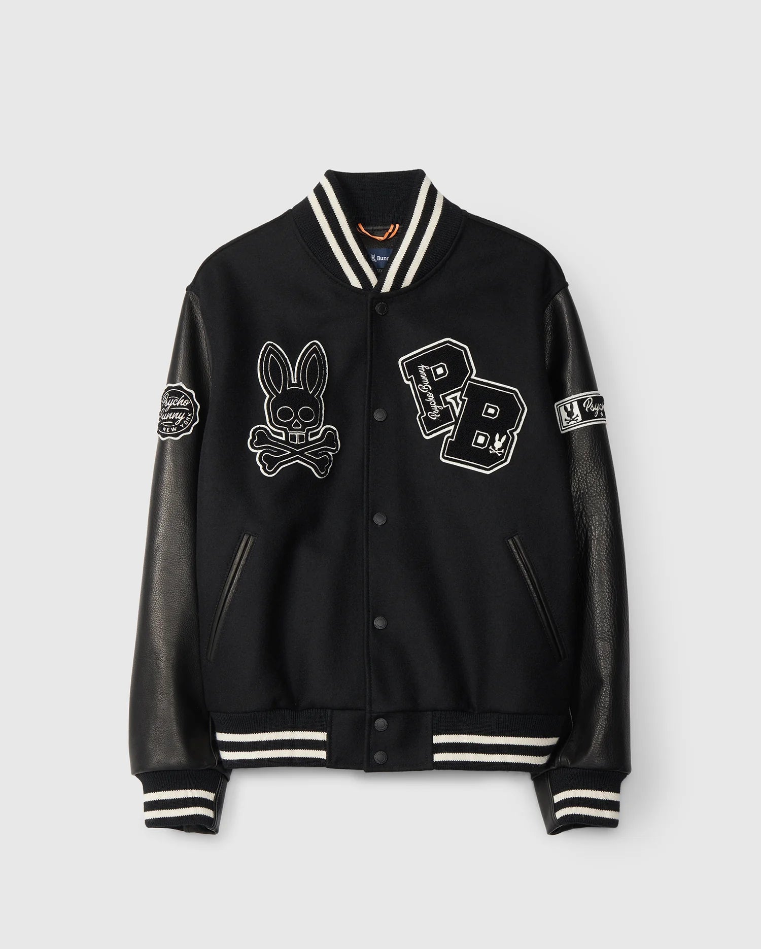 The MENS ROBBINS VARSITY BOMBER JACKET - B6J524Z1OW by Psycho Bunny is a stylish black wool-blend jacket with leather sleeves. It is decorated with embroidered and chenille patches, featuring a skull bunny and "PB" letters, while striped cuffs and collar complete the classic varsity look.