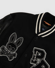 The MENS ROBBINS VARSITY BOMBER JACKET - B6J524Z1OW by Psycho Bunny is a black varsity jacket made from a cozy wool-blend, featuring leather sleeves and a striped collar. It is adorned with an eye-catching skull and crossbones symbol with bunny ears alongside a prominent "P," complete with standout chenille patches for added flair.