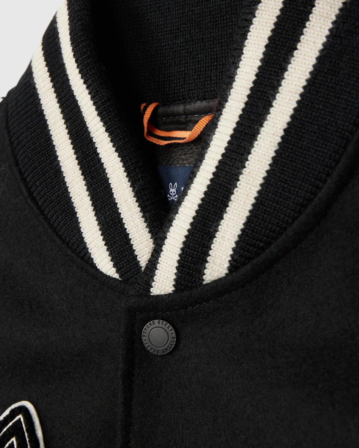A close-up of the Psycho Bunny MENS ROBBINS VARSITY BOMBER JACKET - B6J524Z1OW showcases a striped black and white ribbed collar. The fabric is secured by a single black button, with an orange loop tag partially visible. Sophisticated chenille patches add a touch of athletic elegance to the design.