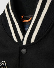 A close-up of the Psycho Bunny MENS ROBBINS VARSITY BOMBER JACKET - B6J524Z1OW showcases a striped black and white ribbed collar. The fabric is secured by a single black button, with an orange loop tag partially visible. Sophisticated chenille patches add a touch of athletic elegance to the design.
