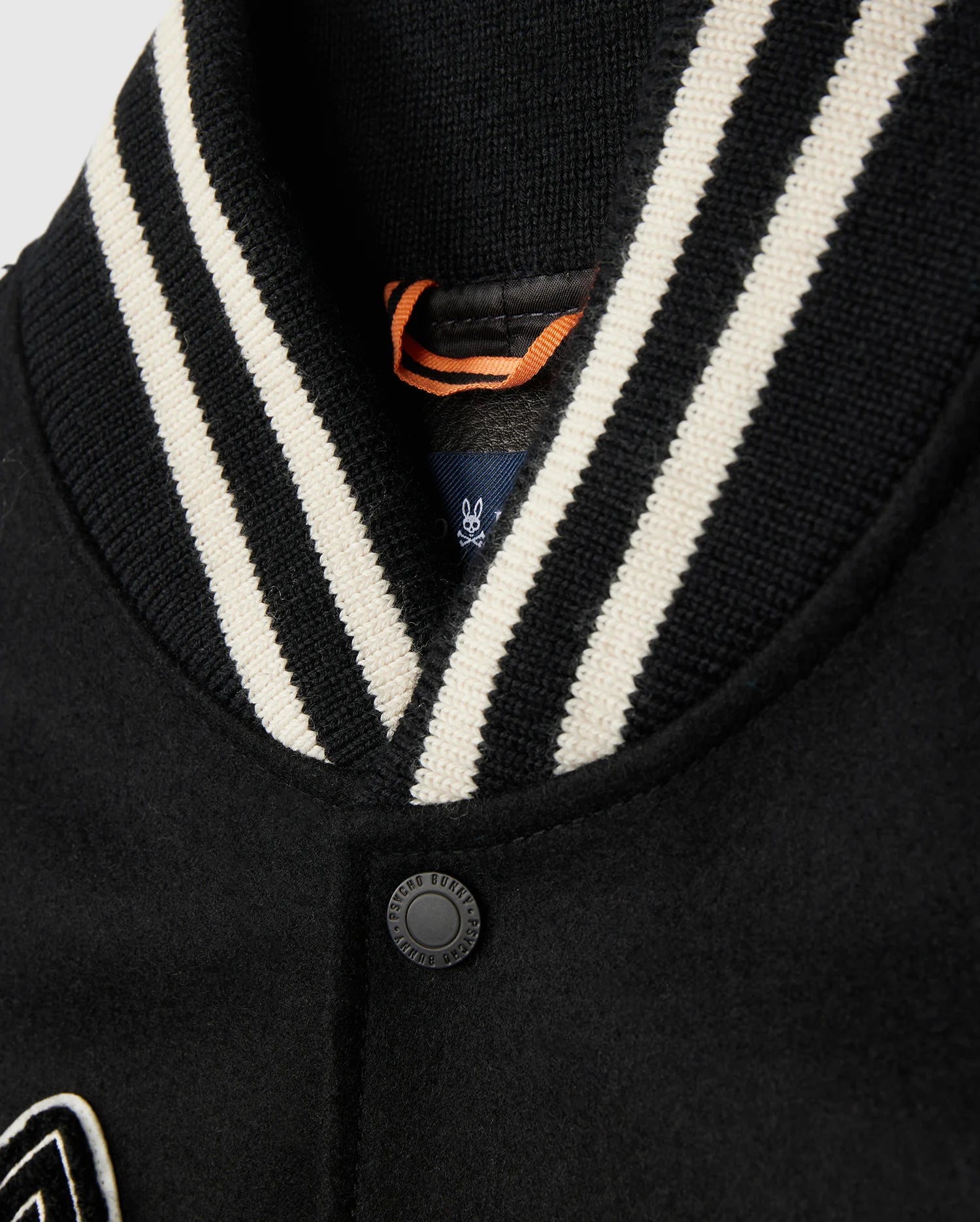 A close-up of the Psycho Bunny MENS ROBBINS VARSITY BOMBER JACKET - B6J524Z1OW showcases a striped black and white ribbed collar. The fabric is secured by a single black button, with an orange loop tag partially visible. Sophisticated chenille patches add a touch of athletic elegance to the design.