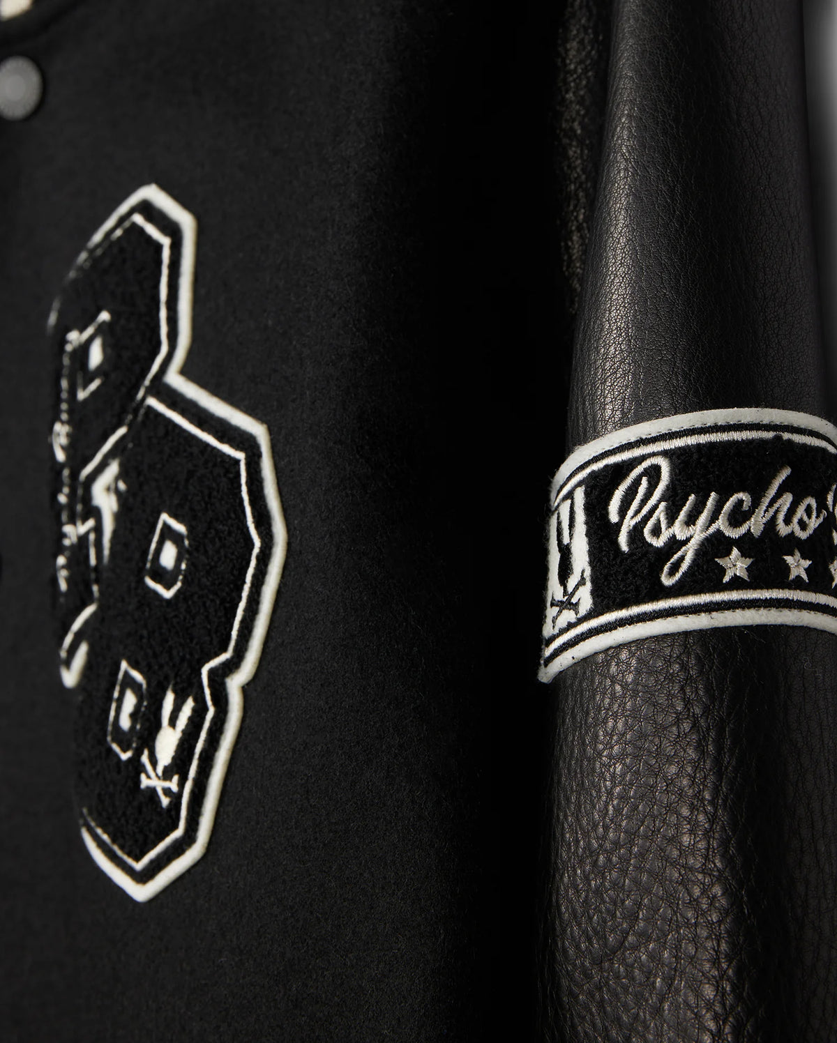 A detailed view of the MENS ROBBINS VARSITY BOMBER JACKET - B6J524Z1OW, crafted by Psycho Bunny. The black leather jacket is embellished with chenille patches, prominently displaying the number "23" with bunny designs. The sleeve features "Psycho" embroidered along with stars. The texture and elaborate stitching are remarkably evident.