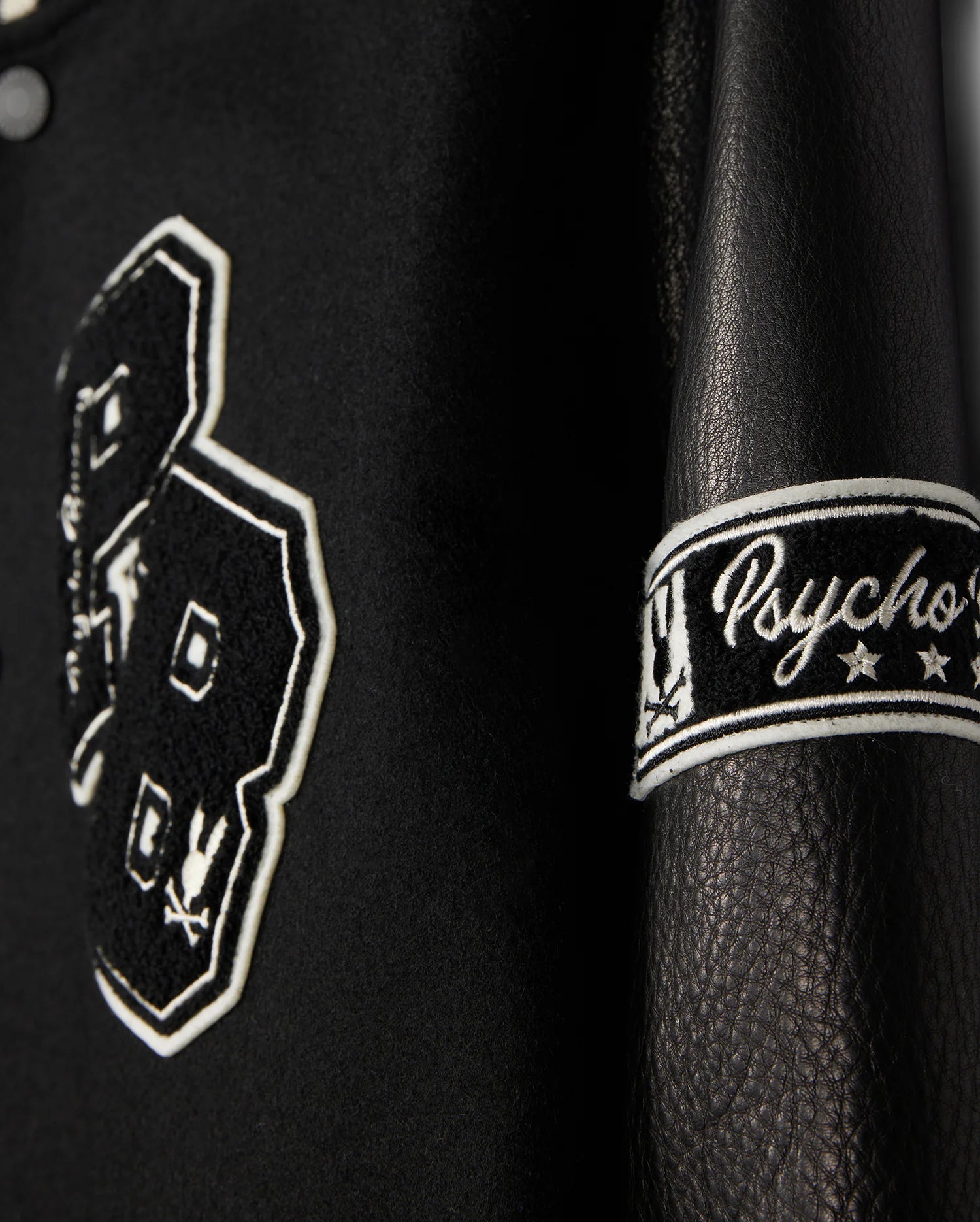 A detailed view of the MENS ROBBINS VARSITY BOMBER JACKET - B6J524Z1OW, crafted by Psycho Bunny. The black leather jacket is embellished with chenille patches, prominently displaying the number "23" with bunny designs. The sleeve features "Psycho" embroidered along with stars. The texture and elaborate stitching are remarkably evident.